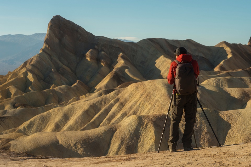 A person in a red jacket, dark pants, and a backpack stands with a tripod on the right side. Bare, tan-colored hills are in the background, with a blue sky.