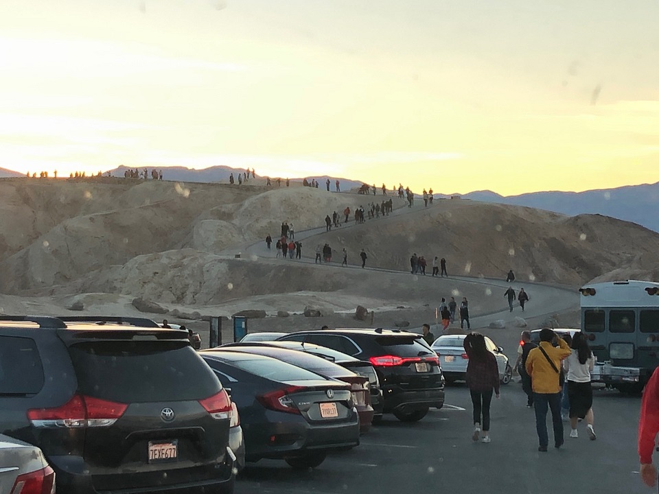 Photo shows a crowded parking lot in the foreground and visitors walking the trail to Zabriskie Point at sunset in the background.