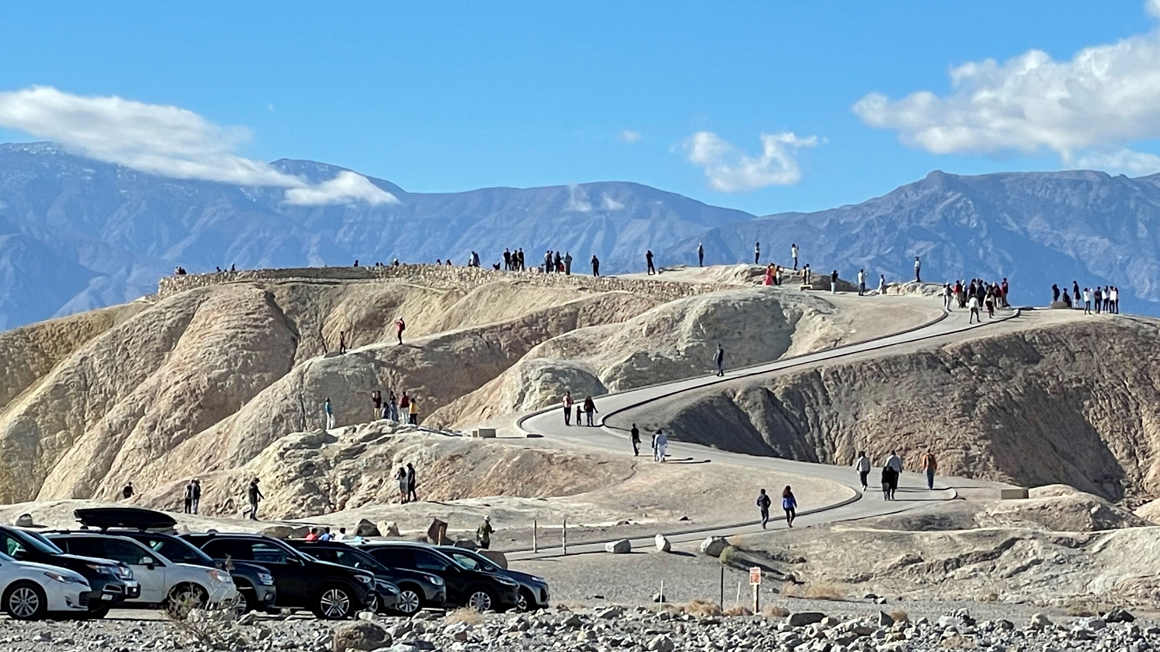 People walk up a winding paved trail from a parking lot to the top of a tan-colored hill. Dark mountains and clouds are in the background.