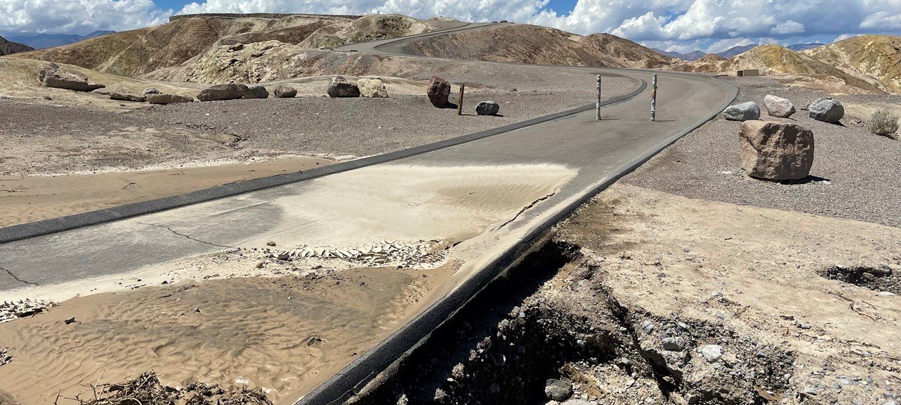 A paved trail leading into badlands is covered in debris and mud with a undercut on the edge of the trail.