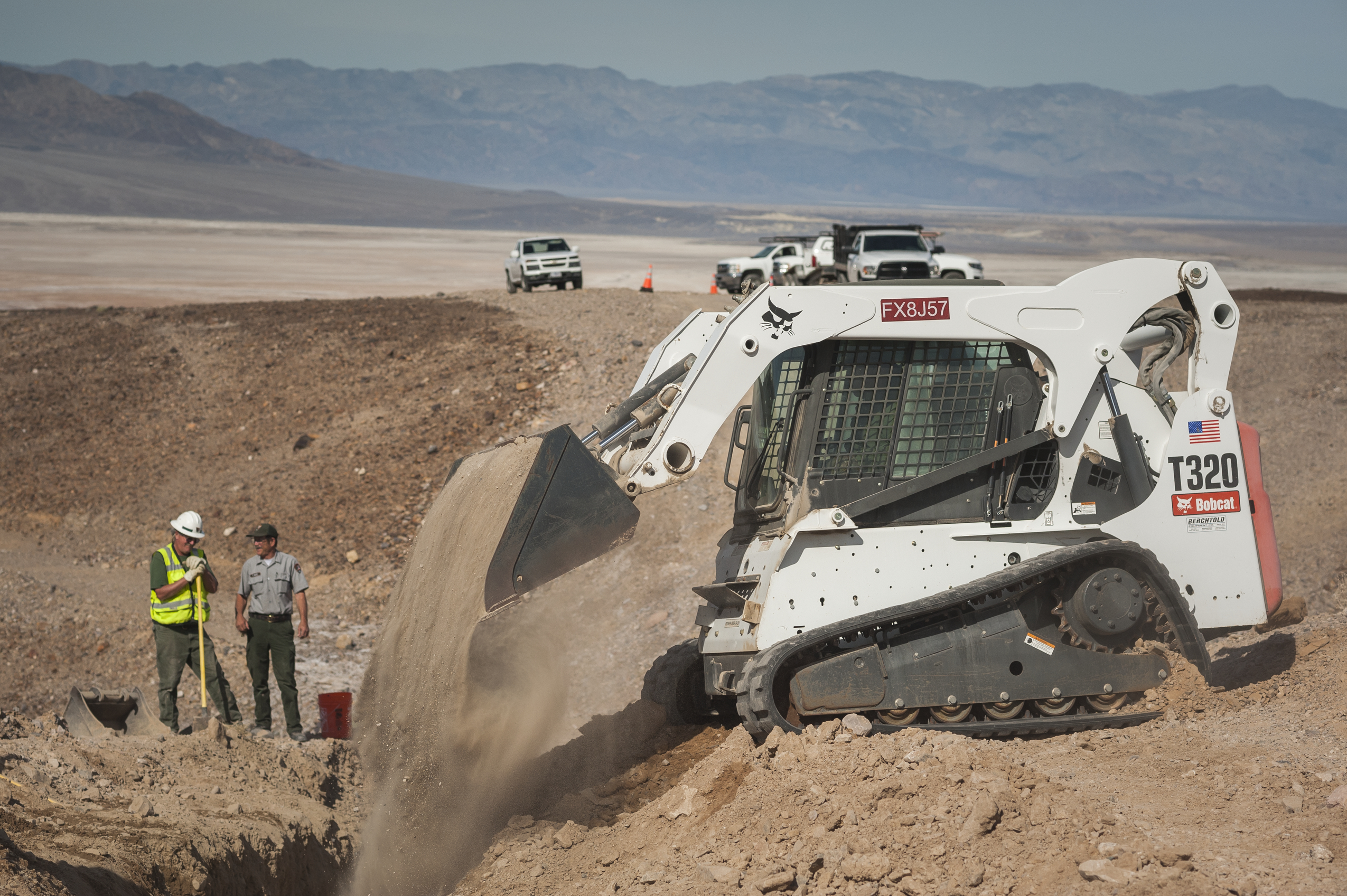 A white T320 Bobcat drops dirt from its bucket into a trench. To people stand next to the trench.