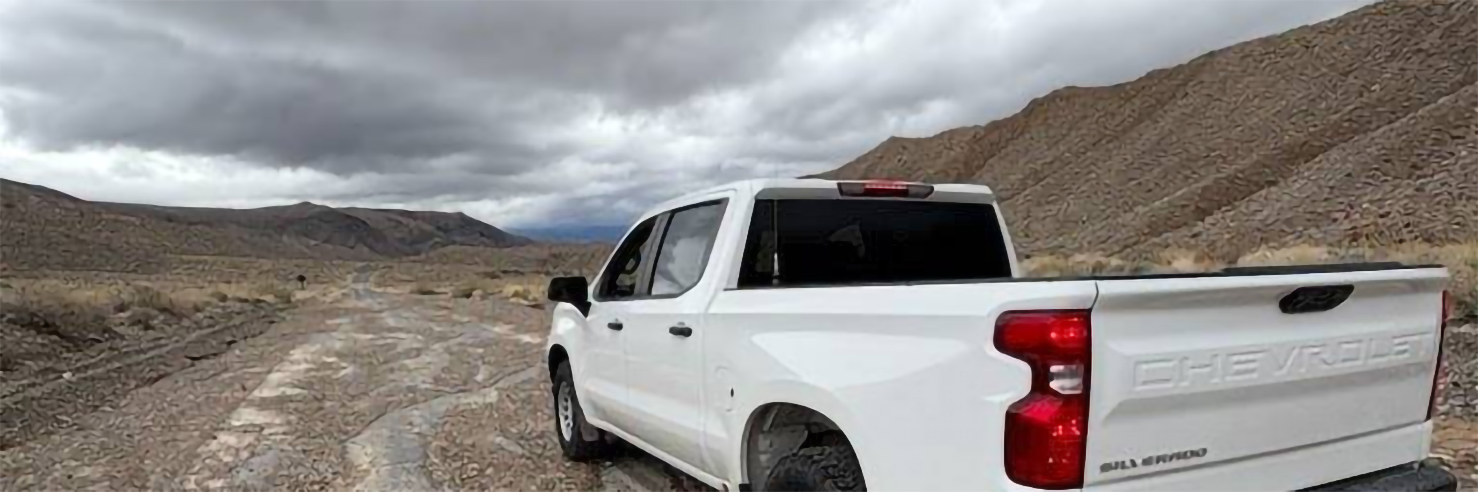 A white pickup truck dominates the right foreground, parked or slowly moving along a rough, rocky dirt track that cuts through a desolate desert valley. Mountain ranges rise up on both sides, beneath a heavy, overcast sky.