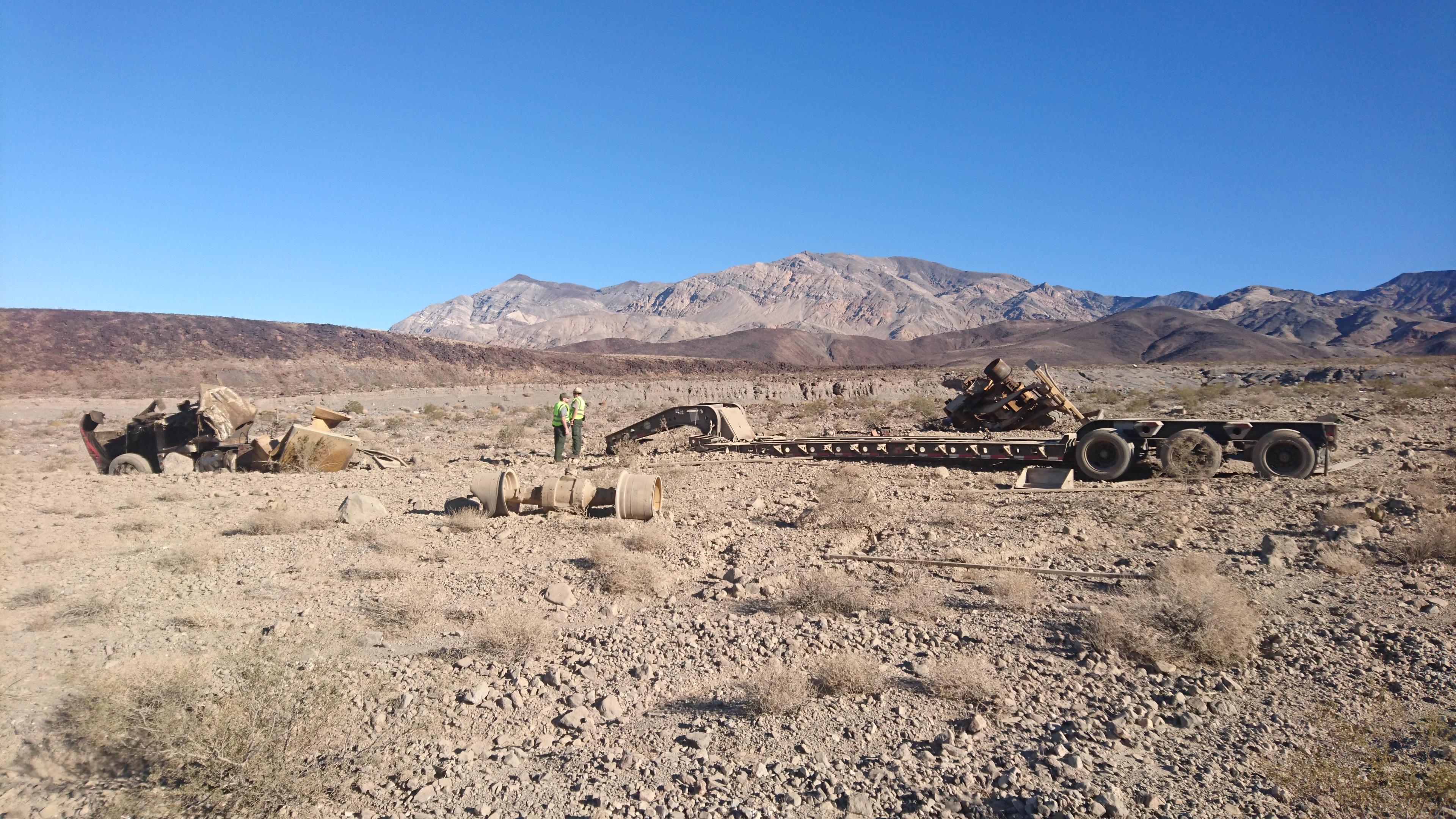 Two employees standing in yellow vests in the distance, with a large tractor trailer overturned next to them. Large brown-hued desert mountains rise in the background.