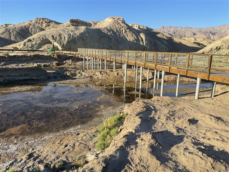 A wooden boardwalk with white supports spans a shallow creek, surrounded by barren desert hills.