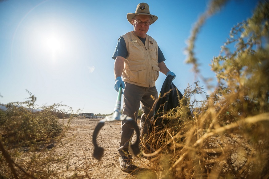 A man wearing a volunteer vest and hat reaches for a piece of litter with a grabber stick.