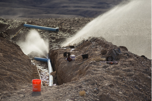 water flowing several feet into the air from a broken blue pipe in a dug out gravel ditch