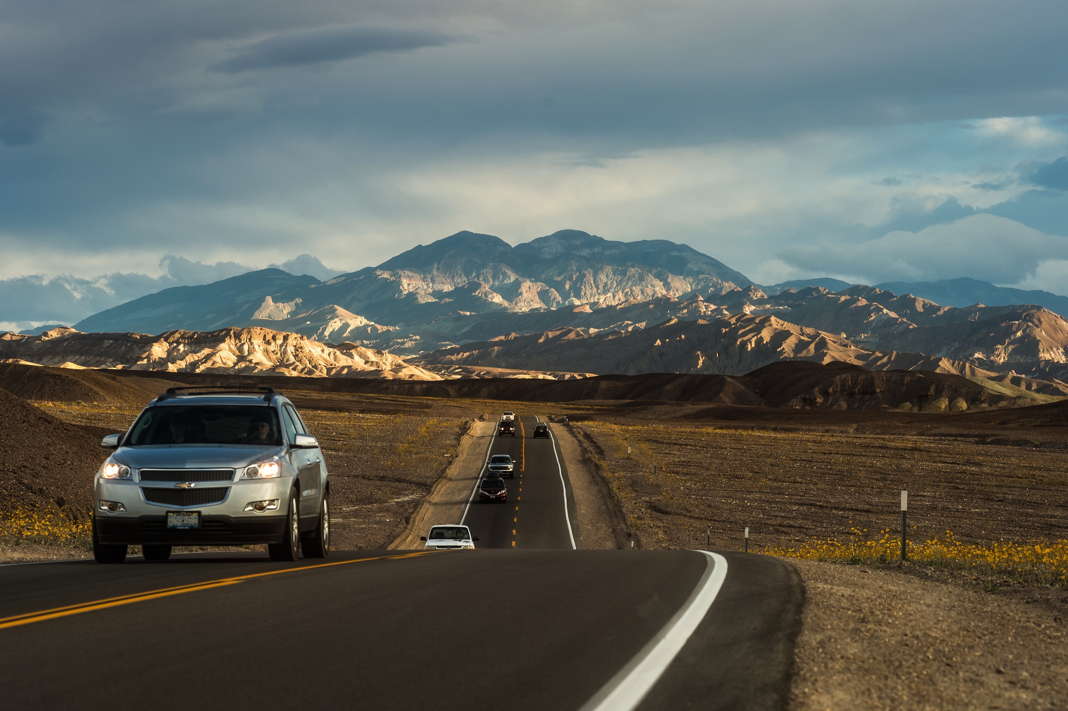 A line of vehicles on a black asphalt road drive with mountains and clouds in the background.