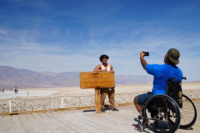 Man in wheelchair taking a photo of his son in front of the Badwater Basin lowest elevation in North America sign