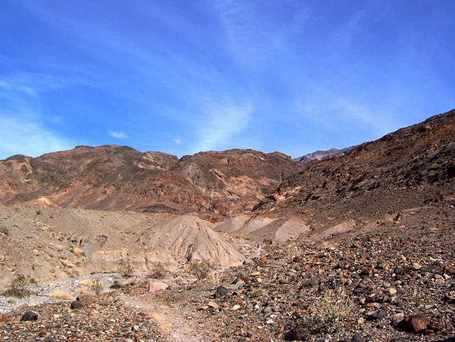 A trail through rocky terrain drops into a dry wash which later turns into a canyon in the reddish brown mountains.