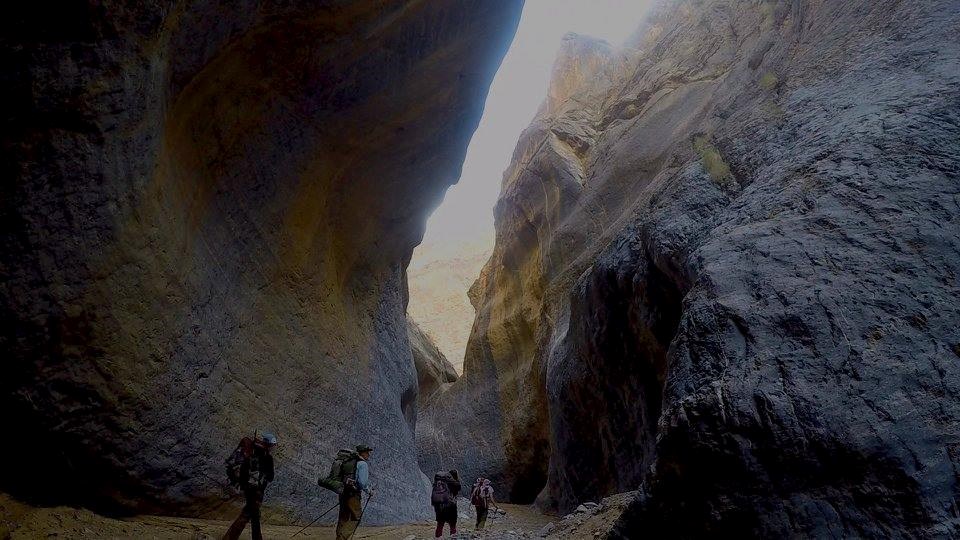 Four backpackers seem tiny as they venture between dark and polished towering canyon walls.