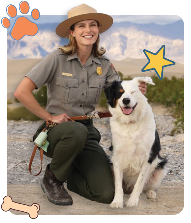 Smiling park ranger kneeling beside a fluffy black-and-white dog on a dirt trail, with mountainous terrain in the distance. The ranger wears a traditional park service uniform and hat, while the dog has one eye closed in a joyful expression.
