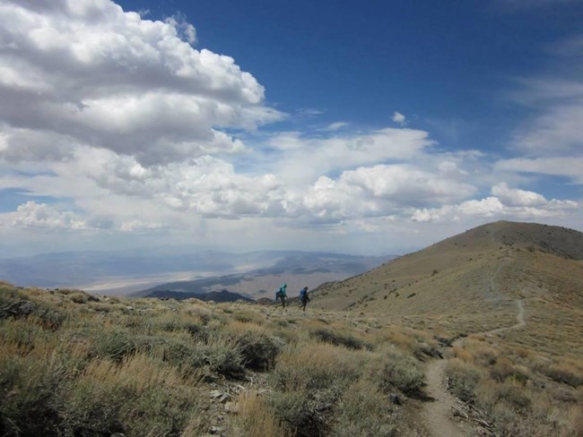 Two hikers descend to an alpine meadow from a narrow ridge.