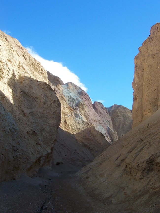 A narrow canyon with red, yellow, and green colored walls beneath a cloudy blue sky.