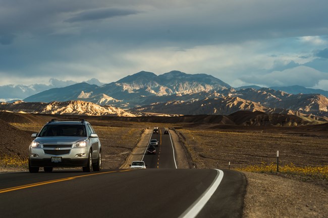 Passenger vehicles drive toward the camera on a paved road with sunlit desert mountains and dark clouds in the background.