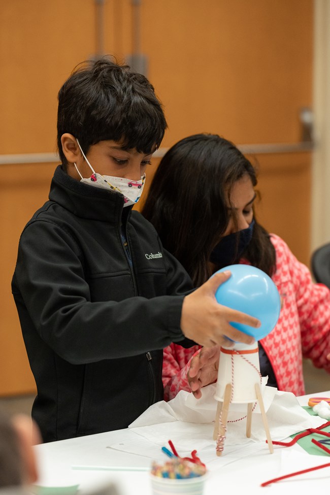 A child attaches a balloon to a paper cup supported by popsicle sticks with a woman in the background.