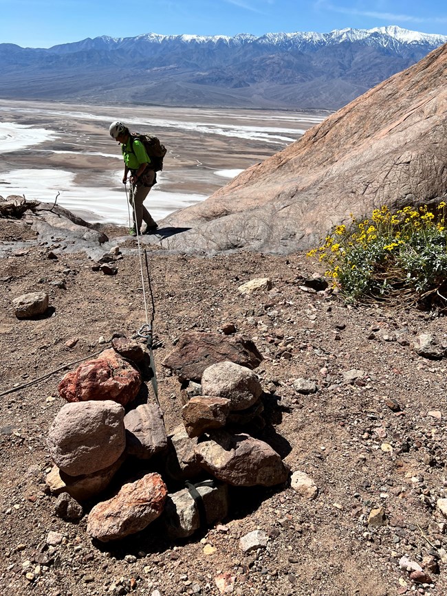 A person stands at the edge of a drop off with a rope attached to a pile of rocks in the foreground.