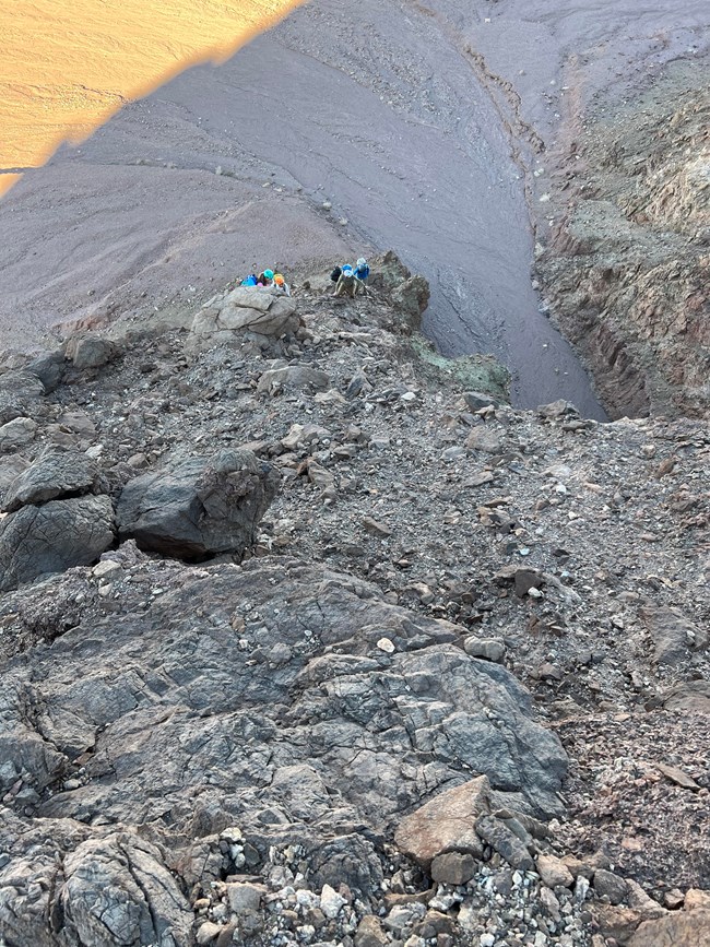 The camera is looking down toward the valley floor. People climb up a very steep rocky slope.