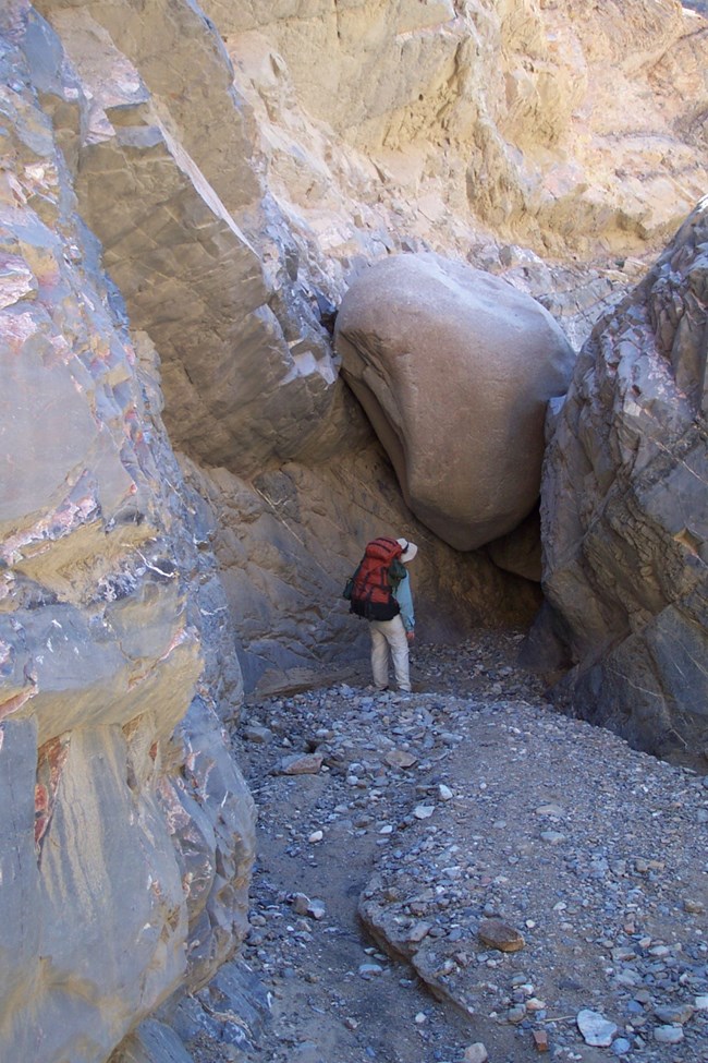Solo backpacker looks up at a large boulder cemented between narrow canyon walls darkened by shadows.
