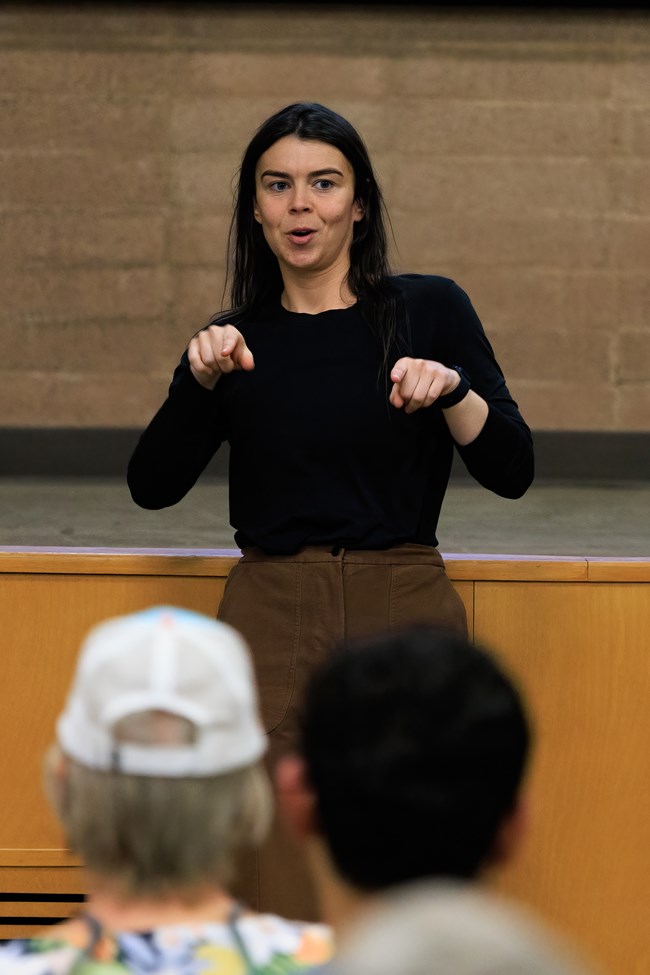 Woman interpreting ASL in front of crowd at auditorium.