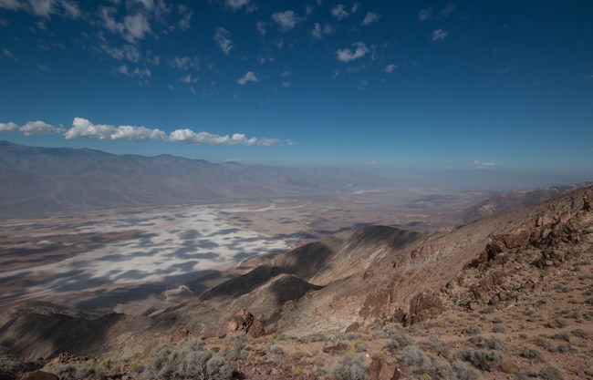 Along a desert ridge, reddish hued mountains drop dramatically down to a flat, white desert valley.