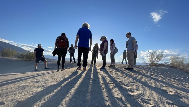 A group of people form a semi-circle around a ranger giving a talk on sand dunes with long shadows falling behind them.