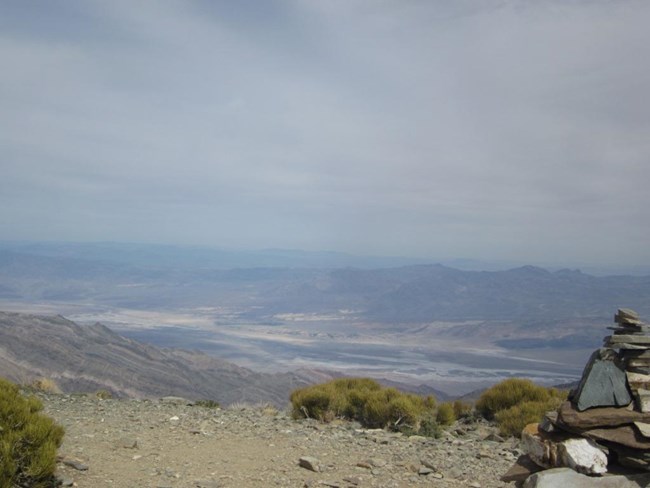 A rock pile marks the summit of Wildrose Peak where the view of the Black Mountains in the distance and Death Valley salt flats lay beneath a cloudy sky.