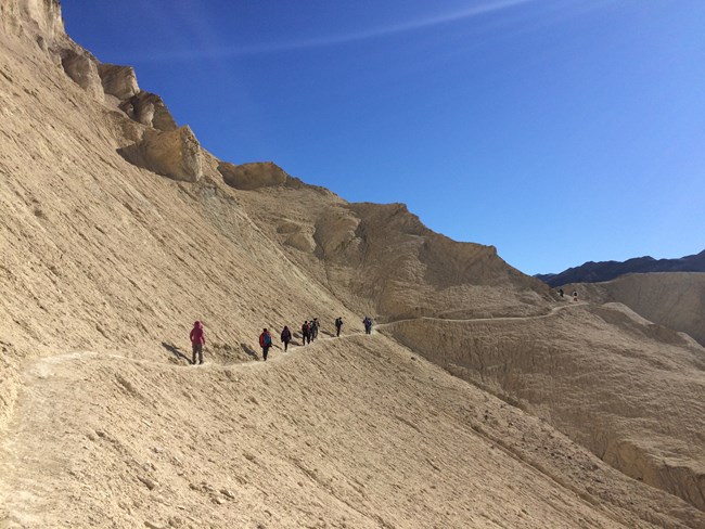 Hikers along a narrow trail below a large golden colored rockface.