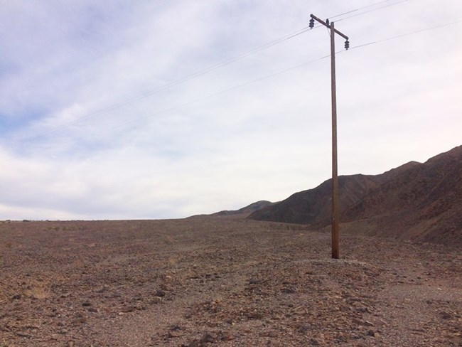 A utility pole stands in a rocky desert wash with mountains in the background.