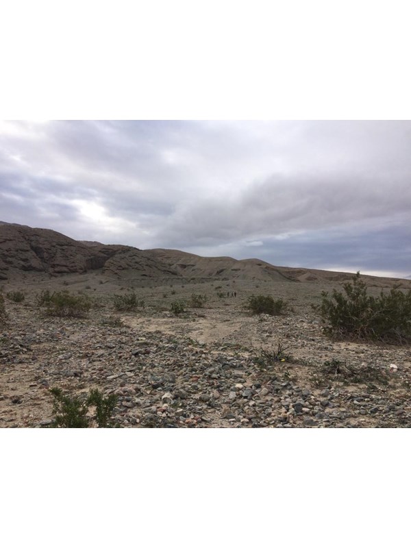 Three hikers walk up an alluvial fan toward the start of a canyon on a grey cloudy day.