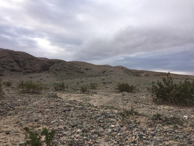 Three hikers walk up an alluvial fan toward the start of a canyon on a grey cloudy day.