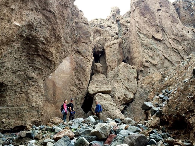 Three hikers stop at a boulder jam in a canyon and face the camera.