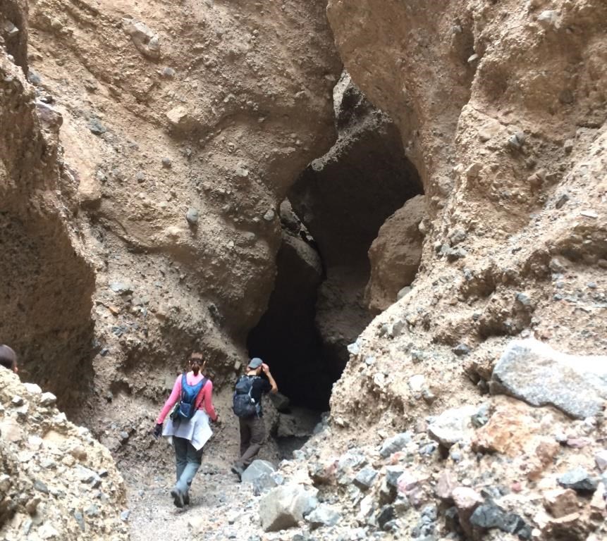 A man and woman hiker head toward a dark slot canyon opening in a canyon wall.