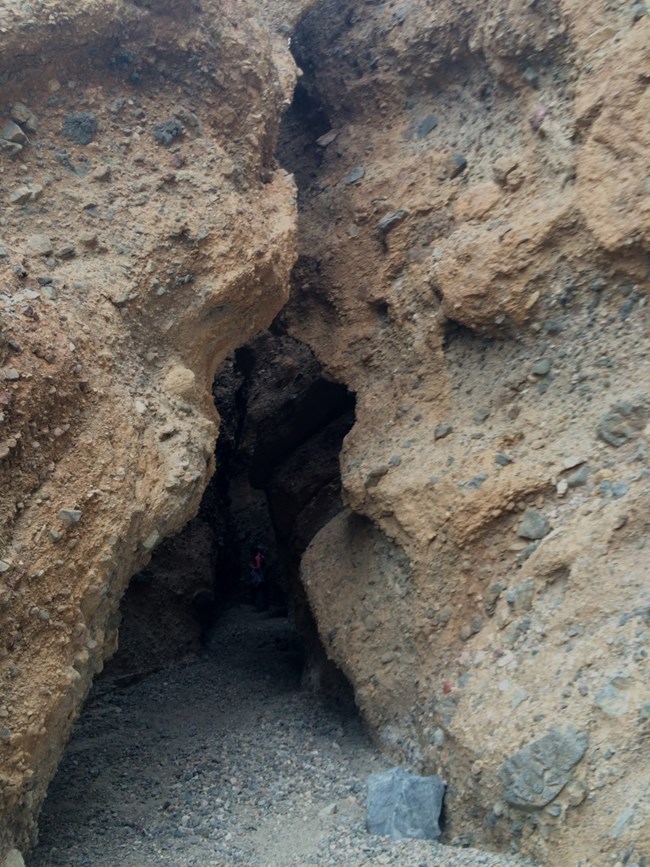 A crack in a canyon wall reveals a dark slot canyon with a hiker exploring inside.