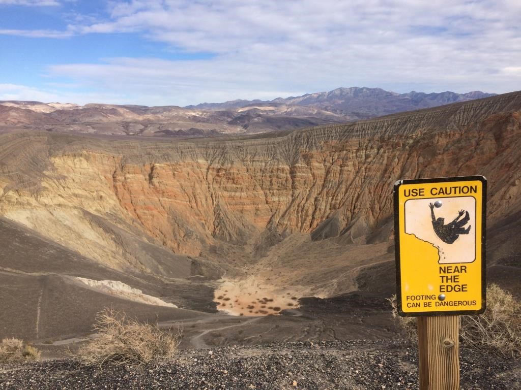 A warning sign "Use caution near edge" with a graphic of someone falling is posted near the edge of a volcanic crater where hikers may travel a loop.