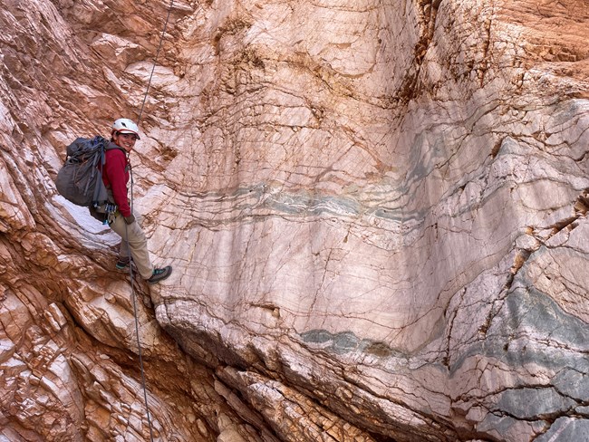 A person wearing a harness hangs from a rope against a polished pink rock wall.