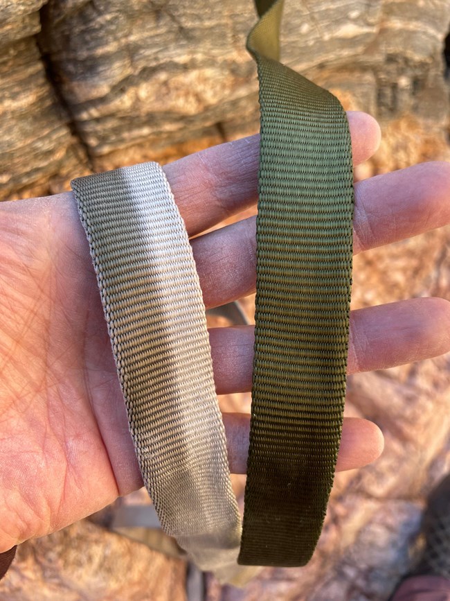 A person holds two pieces of webbing in their hand. On the right is a dark green, new piece. The webbing on the left has been faded by UV light exposure.