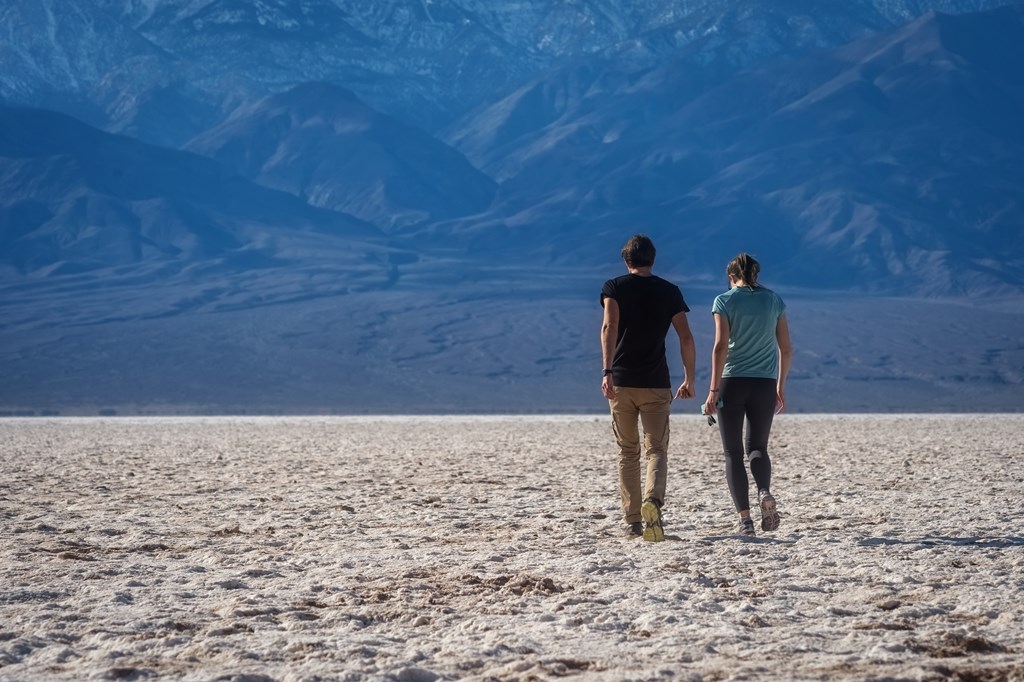 A man and woman walk out onto the white salt flats toward towering mountains in the distance.