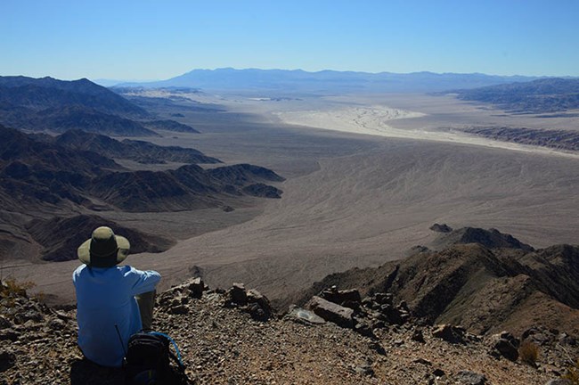 A man in a hat sits on a rugged mountain with a view of a sandy plain.