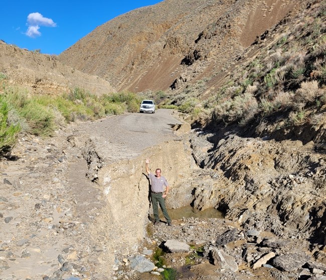 A man in a National Park Service uniform stands on the ground. His arm is extended above his head and does not reach the damaged road surface.