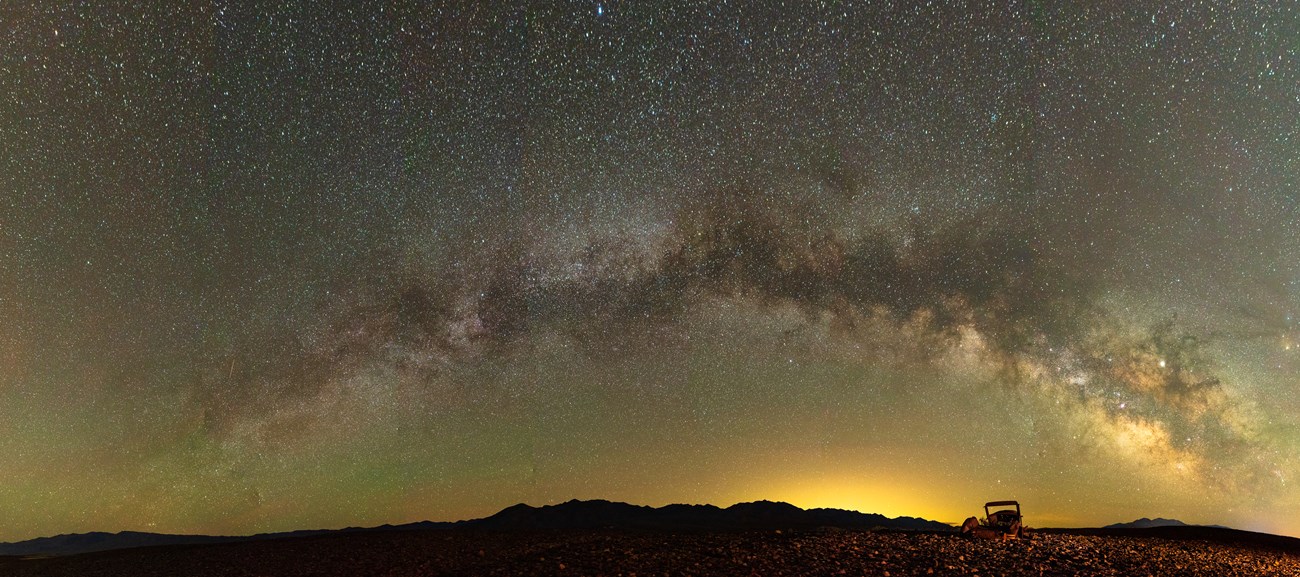 A rocky desert landscape with the remains of a rusty car in the foreground and the Milky Way arching over mountains in the distance in a starry night sky.