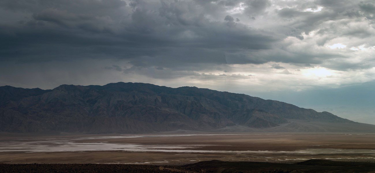 Distant desert mountains, in a cool light, sit under a large dark thunderstorm.