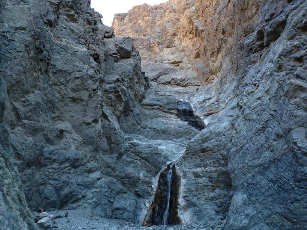 A two-tiered waterfall cascades over polished rock down a narrow canyon as a hiker rests on the ground to the left of the falls with her back against the canyon wall.