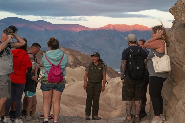 Park Ranger Sarah Carter stands facing a small group of park visitors. Sunrise is just starting to light up the mountains and badlands in the background.