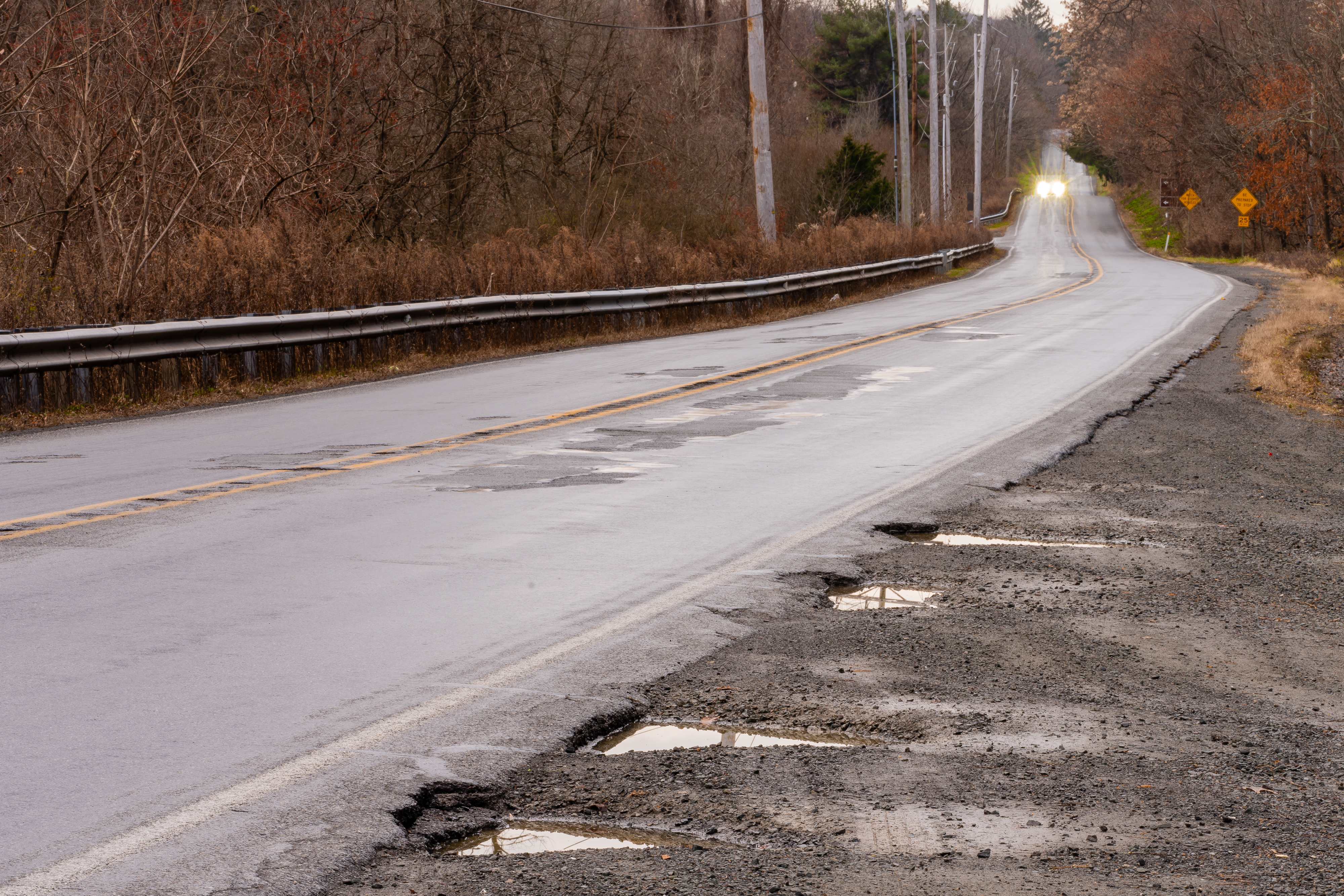 A road with potholes on the right shoulder and the center of the road. An oncoming vehicle's lights are distantly seen in the left lane.