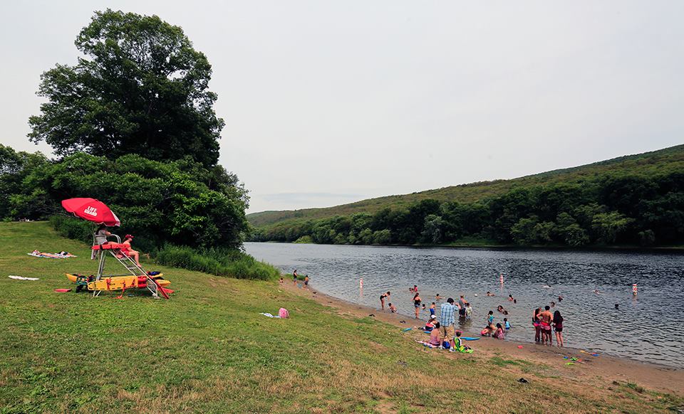 Lifeguards sit under a red umbrella on a tall stand on a grass-covered lawn that extends to the edge of a river while watching about 30 people swimming and wading in the water.