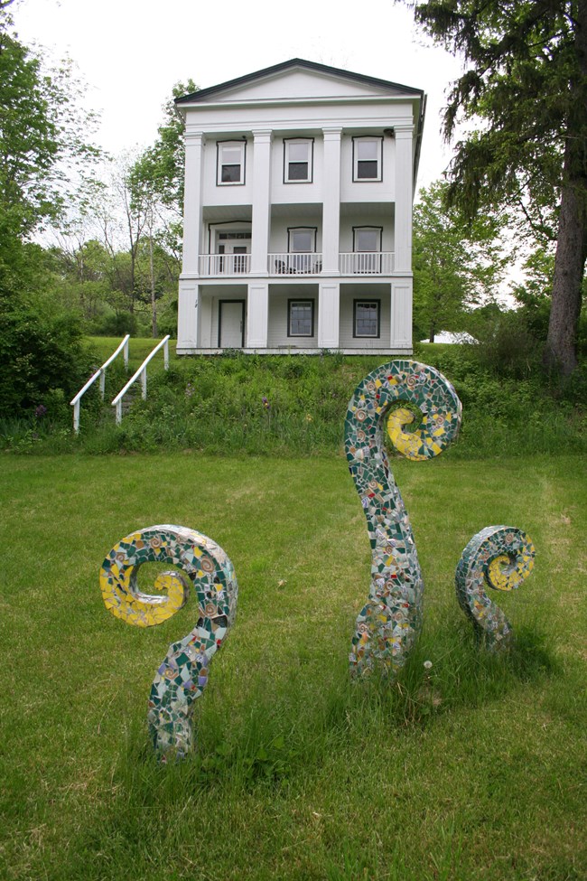 A sculpture rises from the grass in front of a Greek Revival building.