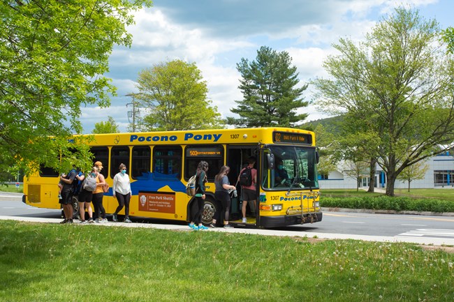 On a sunny day, a large commuter shuttle bus loads passengers. There is a small tree with summer leaves to the left of the bus