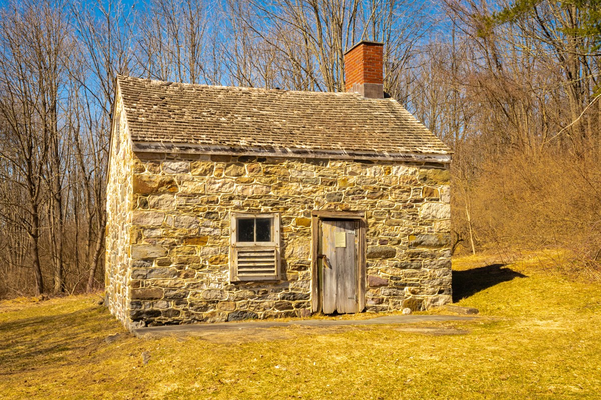 A small stone one room house-like structure with a door to the right and a window to the left of the door. A brick chimney sits on the right side of the peaked roof.