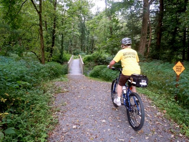 A volunteer in a bright yellow shirt riding a bike on a packed gravel trail.