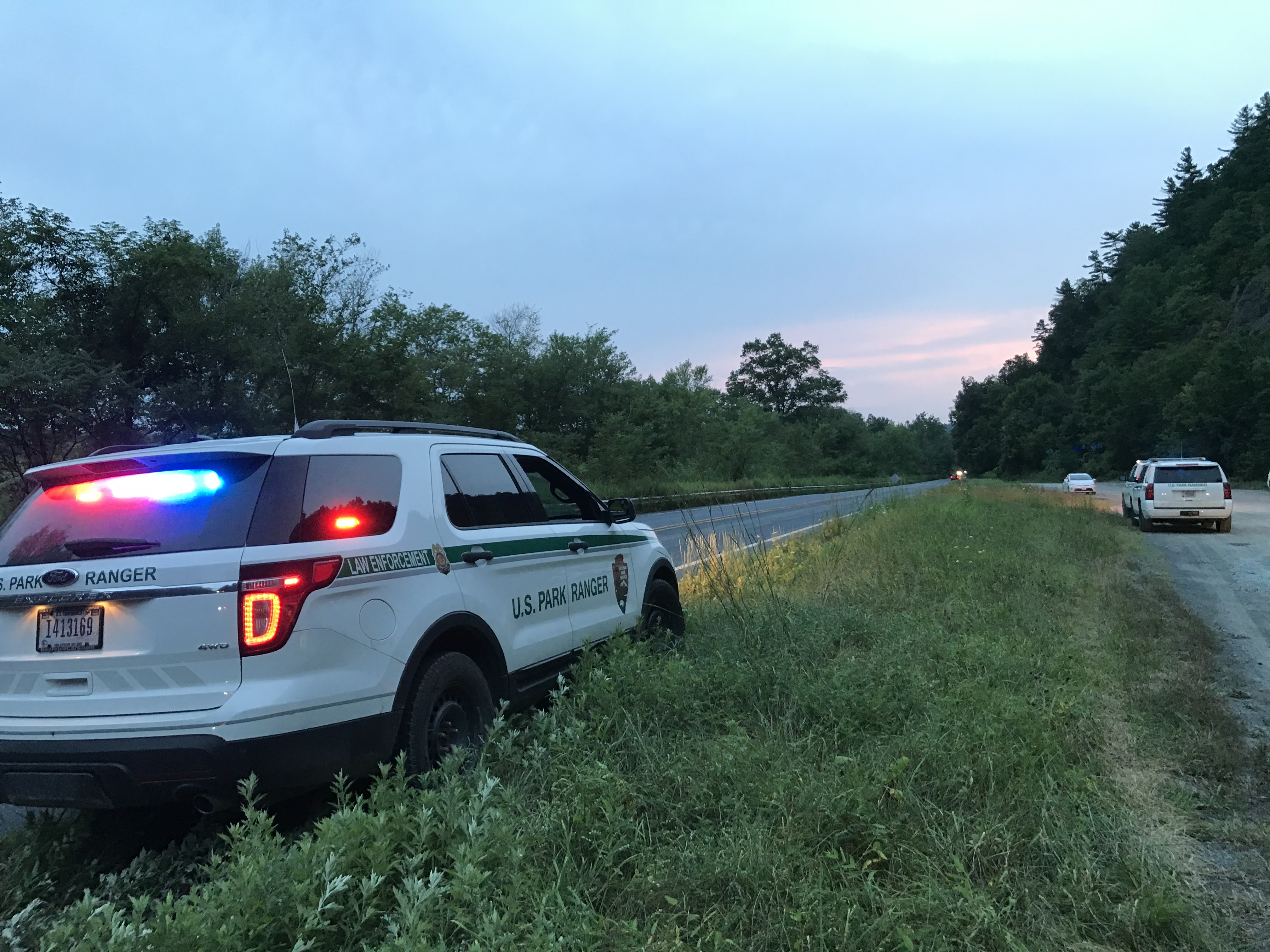A park ranger patrol vehicle with its lights on pulled on the grass next to a road.
